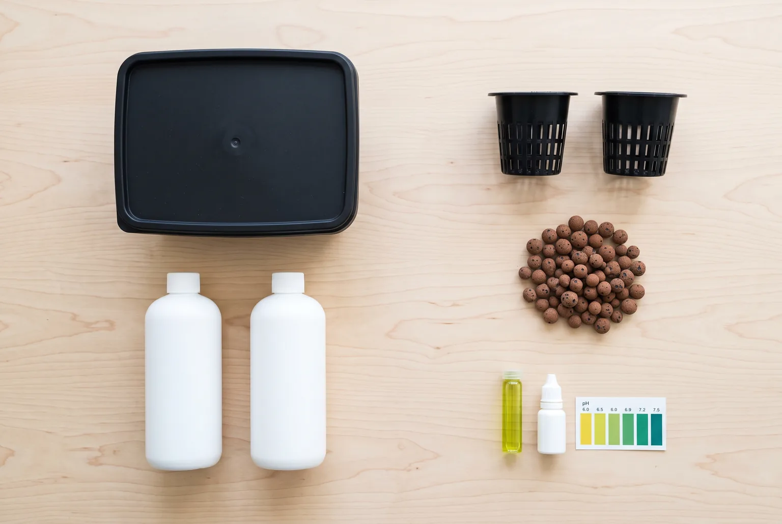 Overhead flat lay of Kratky supplies on a wooden table: an opaque container with lid, two black net cups, a pile of clay pebbles, two nutrient bottles, and a pH test kit